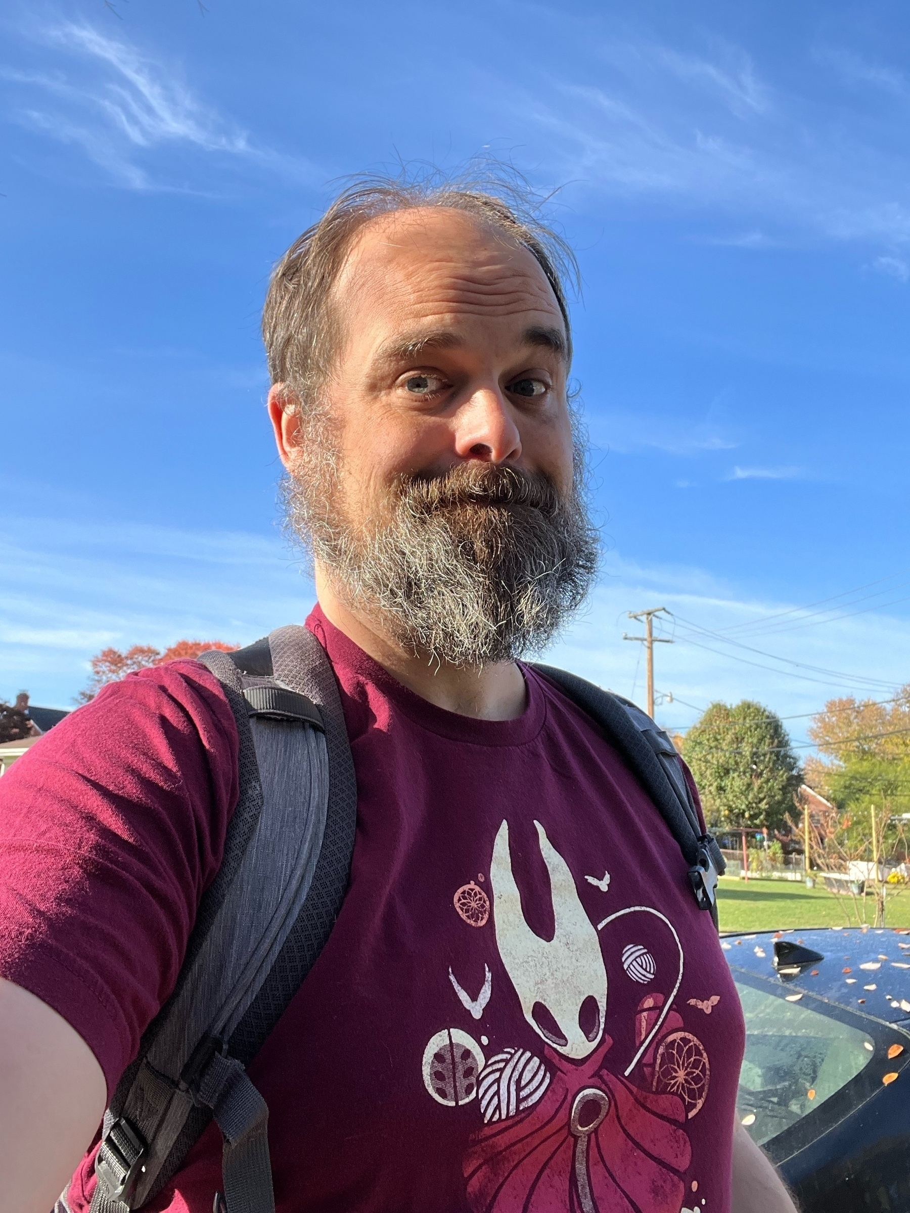 Sam stands in front of a blue sky wearing his Hornet (from Hollow Knight) shirt and his backpack.